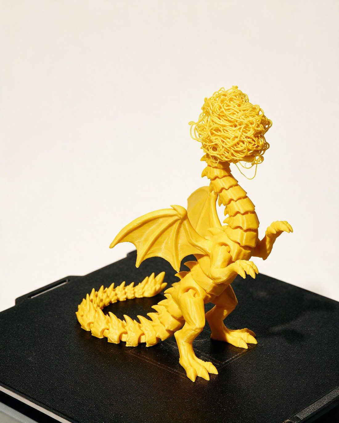 A yellow 3D printed dragon, perfectly formed except for its head, which printed as a tangled nest of filament